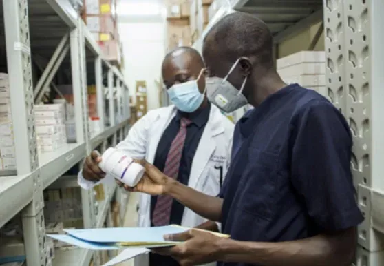 A pharmacist in a white coat and a colleague, both masked, checking a medicine bottle between warehouse shelves