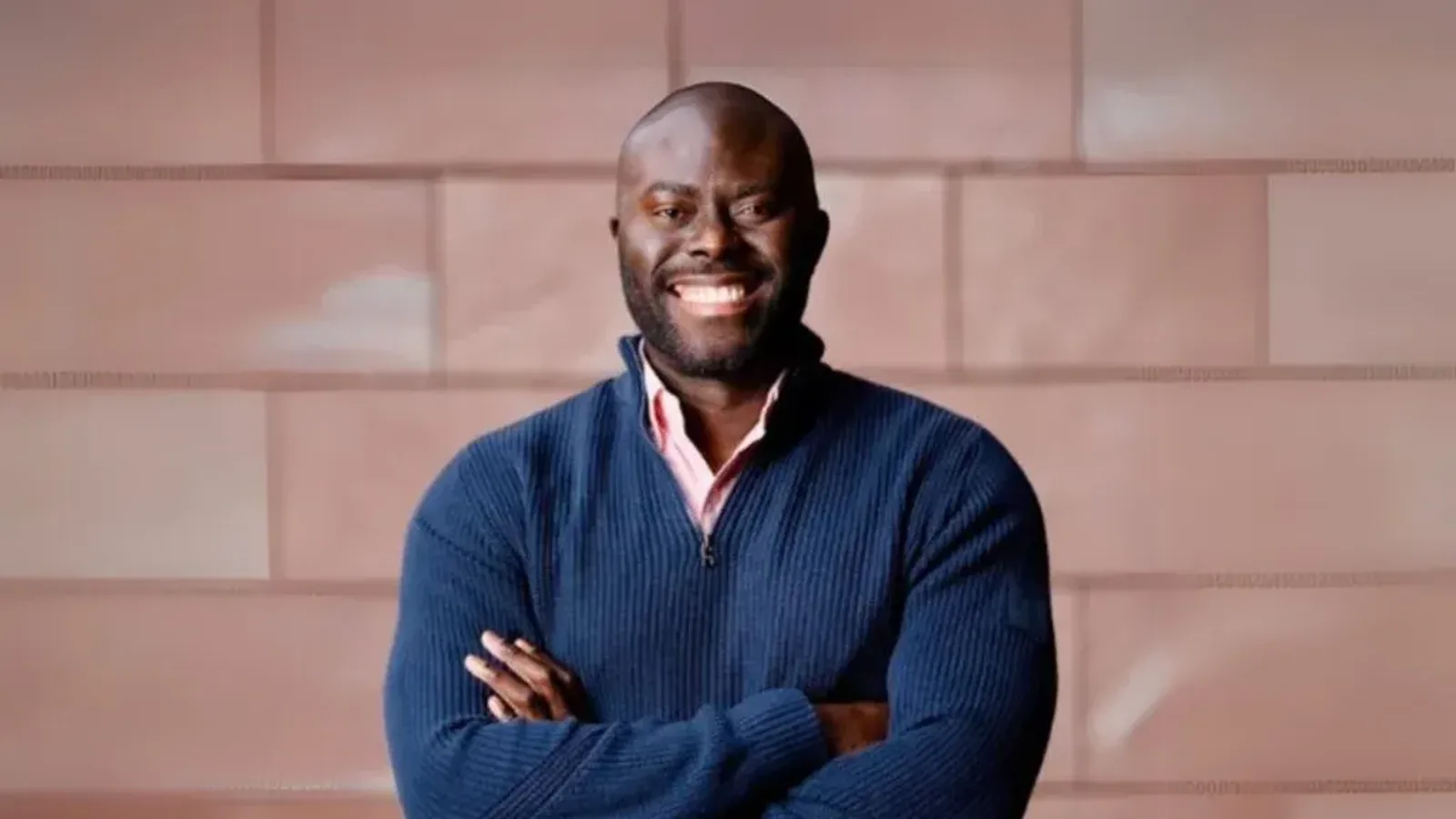 Emmanuel Akpakwu, Founder and CEO of Axmed, smiling with arms folded in front of a sandstone wall