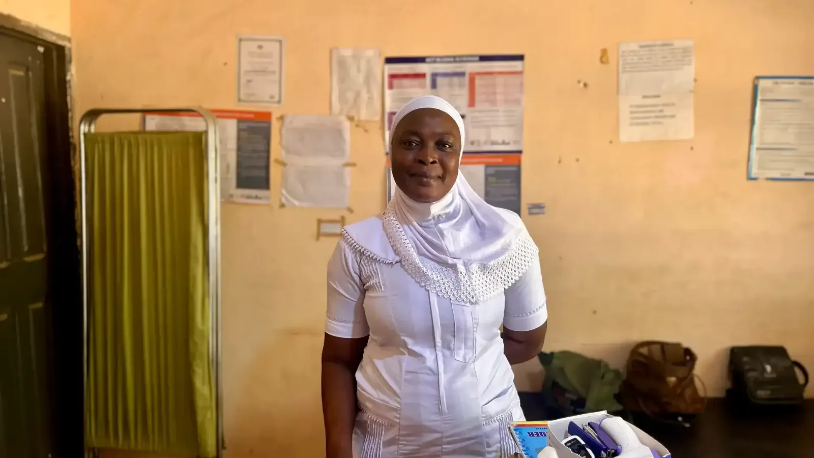 A nurse in a white uniform and headscarf standing in a clinic consulting room in northern Ghana, health posters on the wall behind her